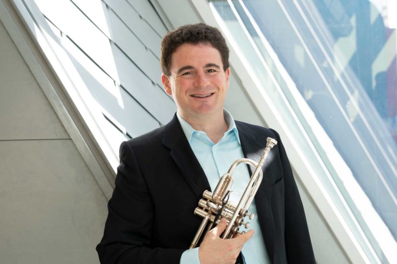 Mike Zonshine wearing a collared shirt and a suit jacket holding a trumpet in front of a window.